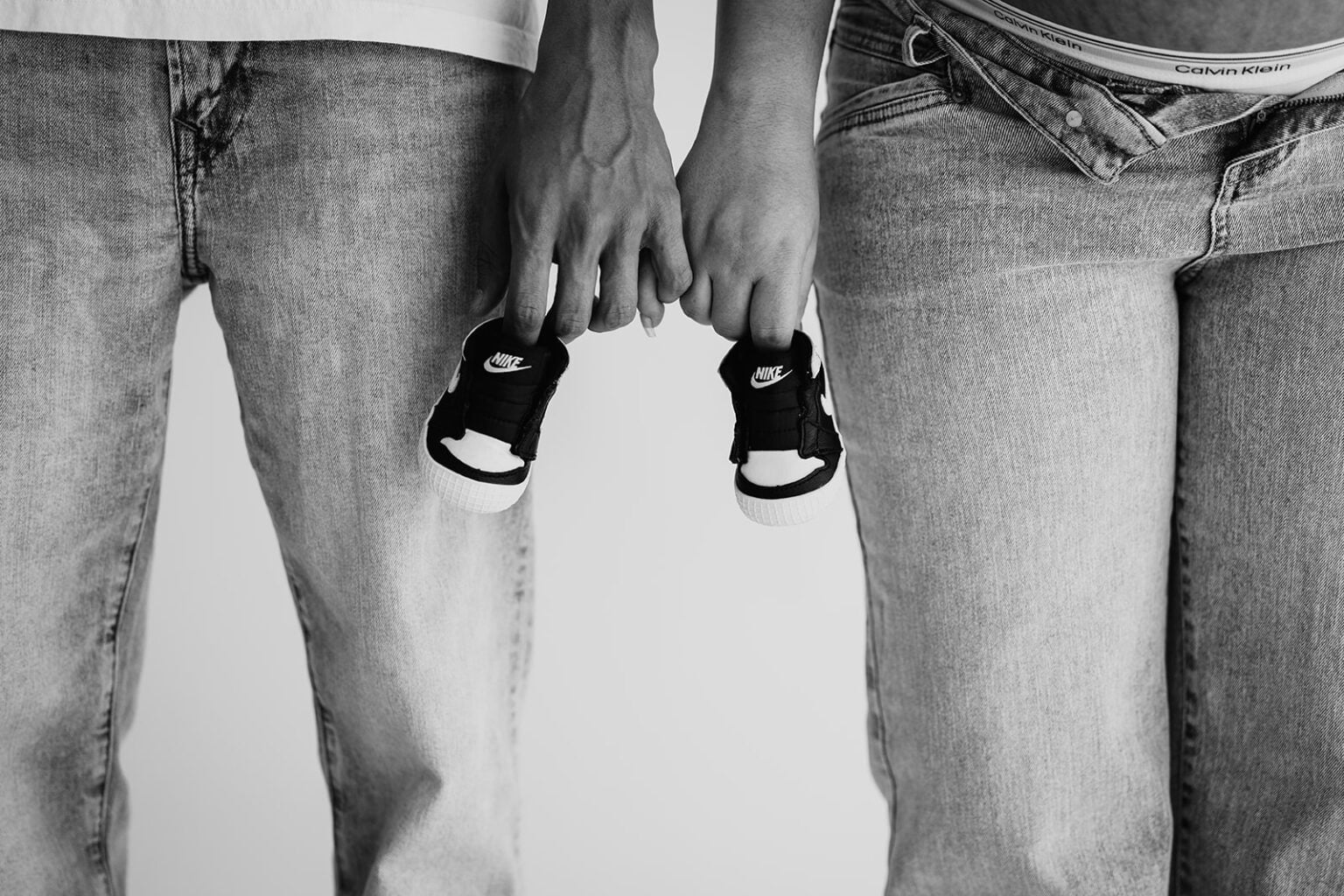 Black-and-white close-up of expecting parents holding hands and a pair of tiny Nike sneakers.