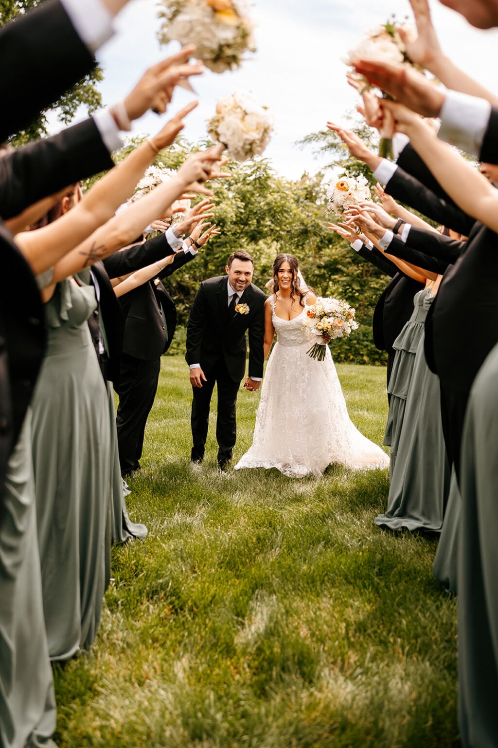 Rachel and Jackson walk hand in hand beneath raised arms and bouquets on the lawn at Mustard Seed Gardens.