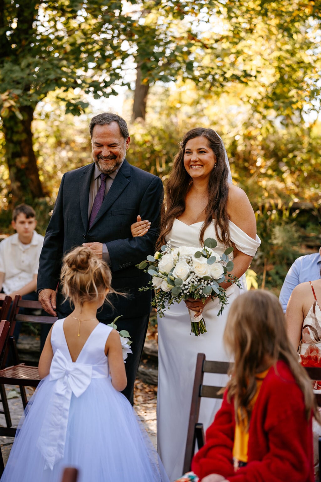 Smiling bride carrying a white bouquet walks down the outdoor ceremony aisle at Alfred Caldwell Lily Pool in Chicago.