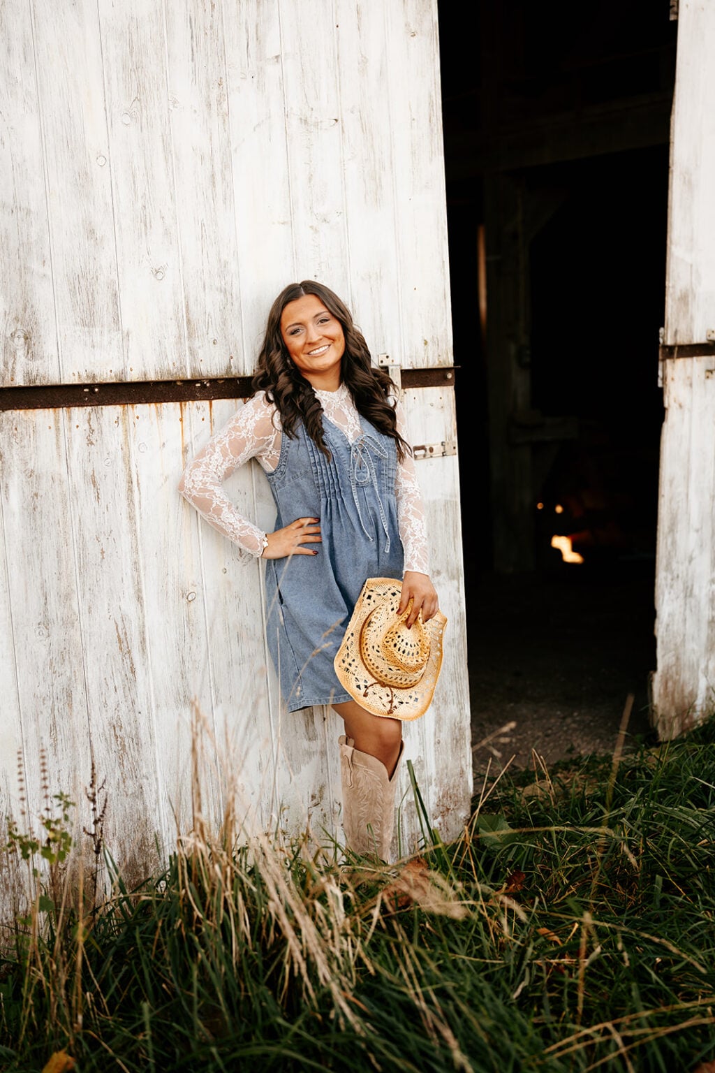 Senior girl in a denim dress and cowboy boots posing beside a white rustic barn.