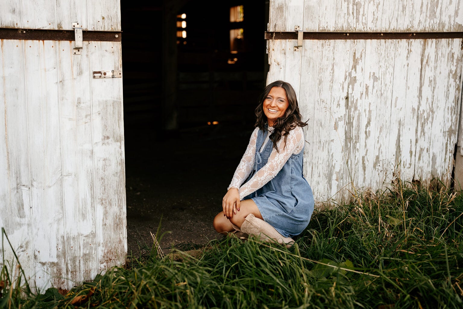 Senior girl in a denim dress and cowboy boots kneeling beside the open doors of a rustic white barn.