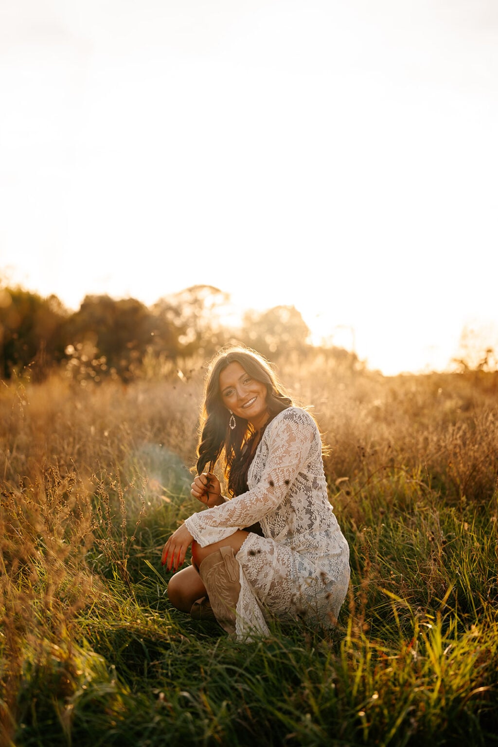 Senior girl wearing a white lace dress and cowboy boots in a sunlit wildflower field.